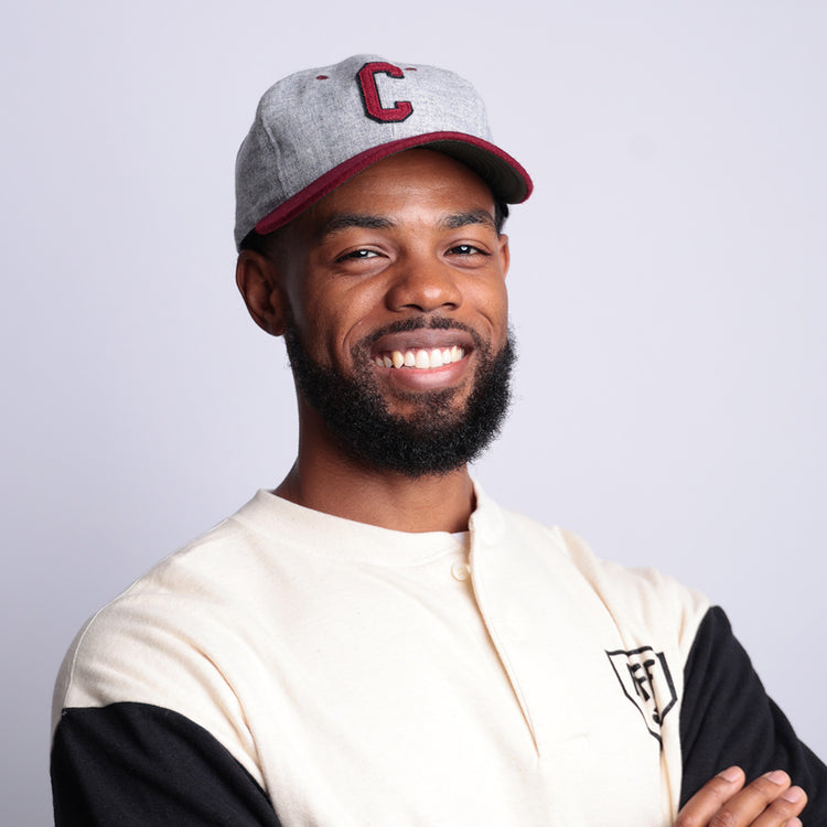 A smiling bearded man wears an EFF University of South Carolina 1949 Vintage Ballcap in light gray with a red C, paired with a cream and black shirt, standing against a plain light background with crossed arms.