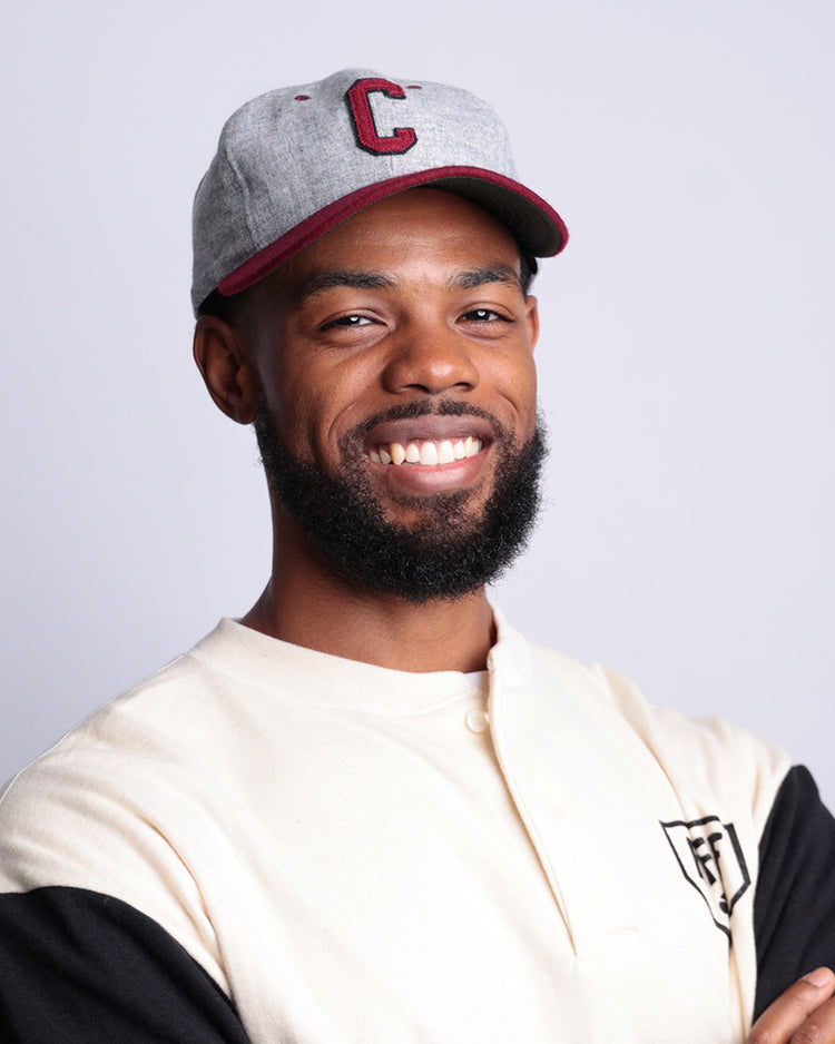 A smiling bearded man wears an EFF University of South Carolina 1949 Vintage Ballcap in light gray with a red C, paired with a cream and black shirt, standing against a plain light background with crossed arms.