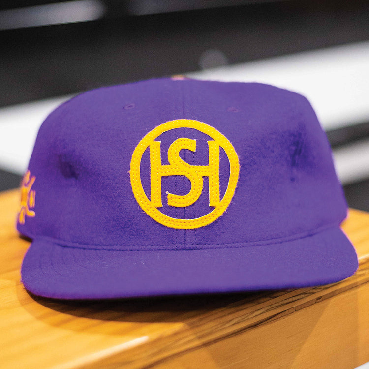 The EFF Spokane Canaries 1917 Vintage Ballcap is a purple wool cap featuring a yellow circular S-H logo on the front and an adjustable leather strap, inspired by the Pacific Coast Hockey Association. Shown resting on a wooden surface.
