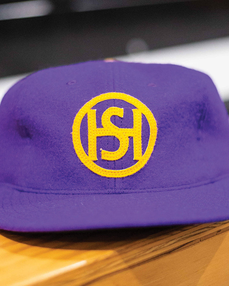 The EFF Spokane Canaries 1917 Vintage Ballcap is a purple wool cap featuring a yellow circular S-H logo on the front and an adjustable leather strap, inspired by the Pacific Coast Hockey Association. Shown resting on a wooden surface.