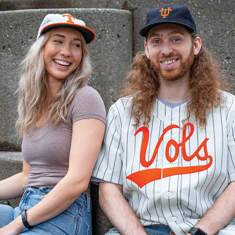 Two people sit on steps, smiling. The woman wears jeans, a gray tee, and the Ebbets Field Flannels University of Tennessee 1962 Vintage Ballcap with an orange T. The man, with long red hair, wears a black vintage cap and a white Vols baseball jersey.