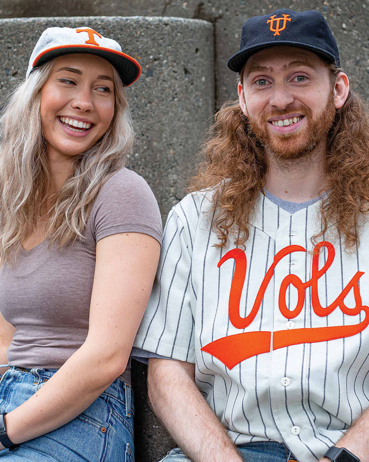 Two people sit on steps, smiling. The woman wears jeans, a gray tee, and the Ebbets Field Flannels University of Tennessee 1962 Vintage Ballcap with an orange T. The man, with long red hair, wears a black vintage cap and a white Vols baseball jersey.