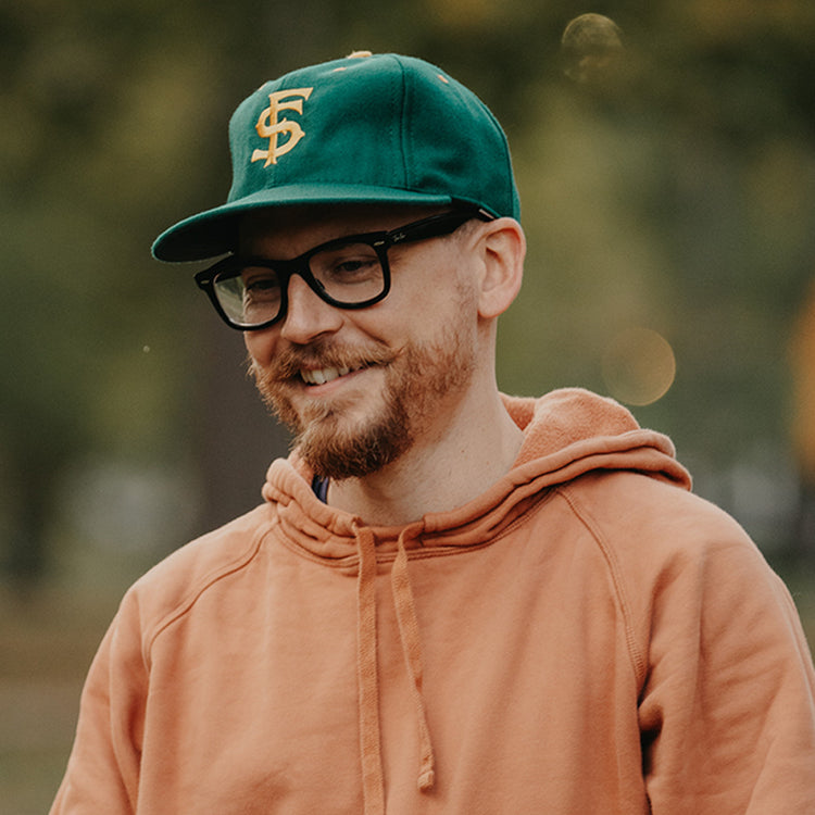 A smiling man with a reddish beard and glasses wears an orange hoodie and the EFF University of San Francisco 1950 Vintage Ballcap, standing outdoors with blurred trees in the background.