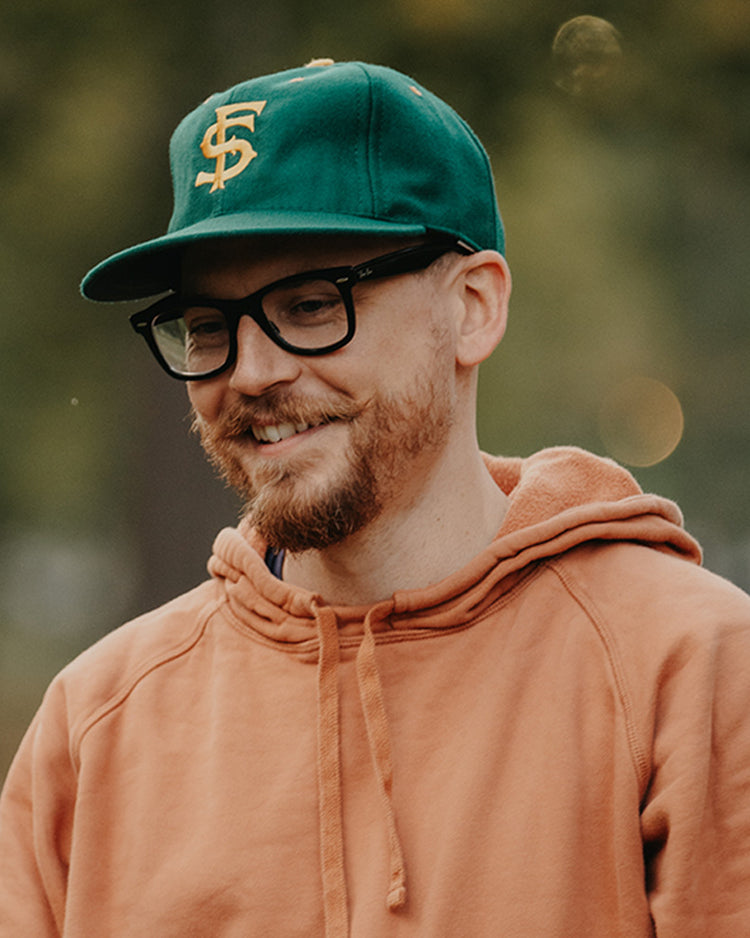 A smiling man with a reddish beard and glasses wears an orange hoodie and the EFF University of San Francisco 1950 Vintage Ballcap, standing outdoors with blurred trees in the background.