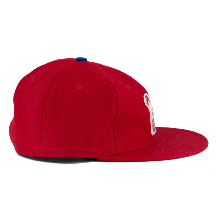 The EFF Wembley Lions 1957 Vintage Ballcap is a hand-sewn red wool cap with a flat brim, a small blue top button, and a partial white and red embroidered logo on the front panel, shown from the side.