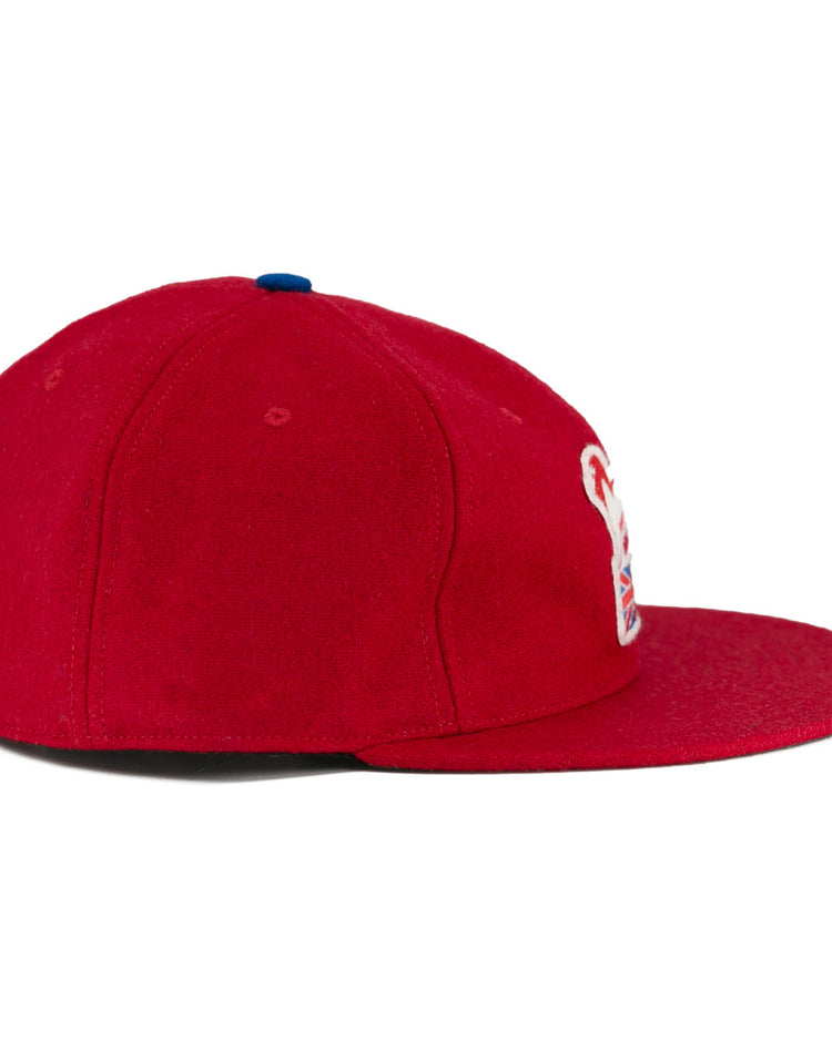 The EFF Wembley Lions 1957 Vintage Ballcap is a hand-sewn red wool cap with a flat brim, a small blue top button, and a partial white and red embroidered logo on the front panel, shown from the side.
