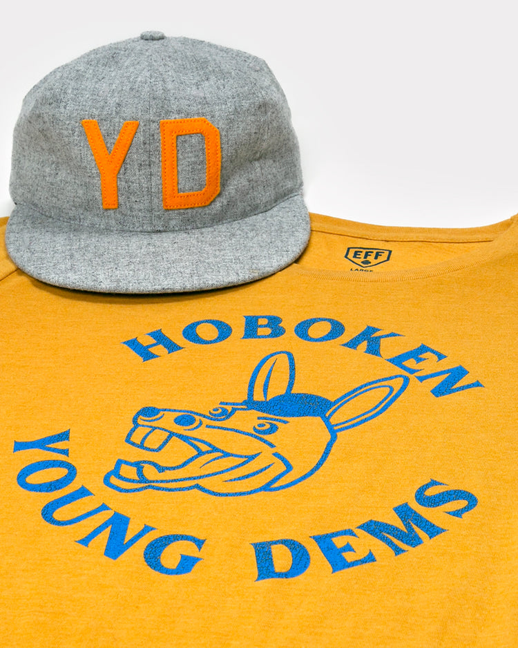 A gray cap with orange YD sits on a yellow Hoboken Young Democrats T-Shirt by Ebbets Field Flannels, featuring a blue cartoon donkey and Hoboken Young Dems in a circular design.