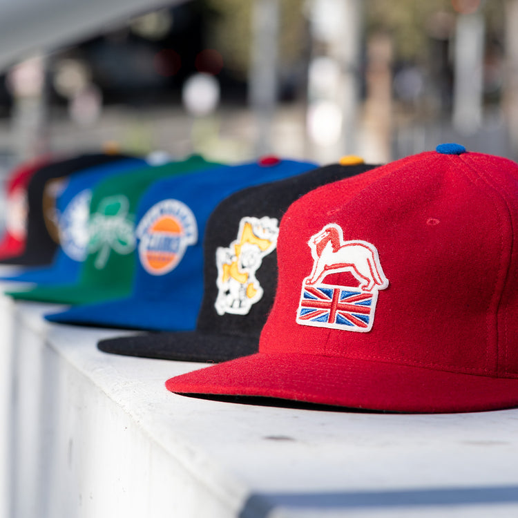 A row of colorful baseball caps is displayed outdoors, highlighting the EFF Wembley Lions 1957 Vintage Ballcap—a hand-sewn wool red cap with a white horse and British flag patch—reminiscent of classic vintage styles.