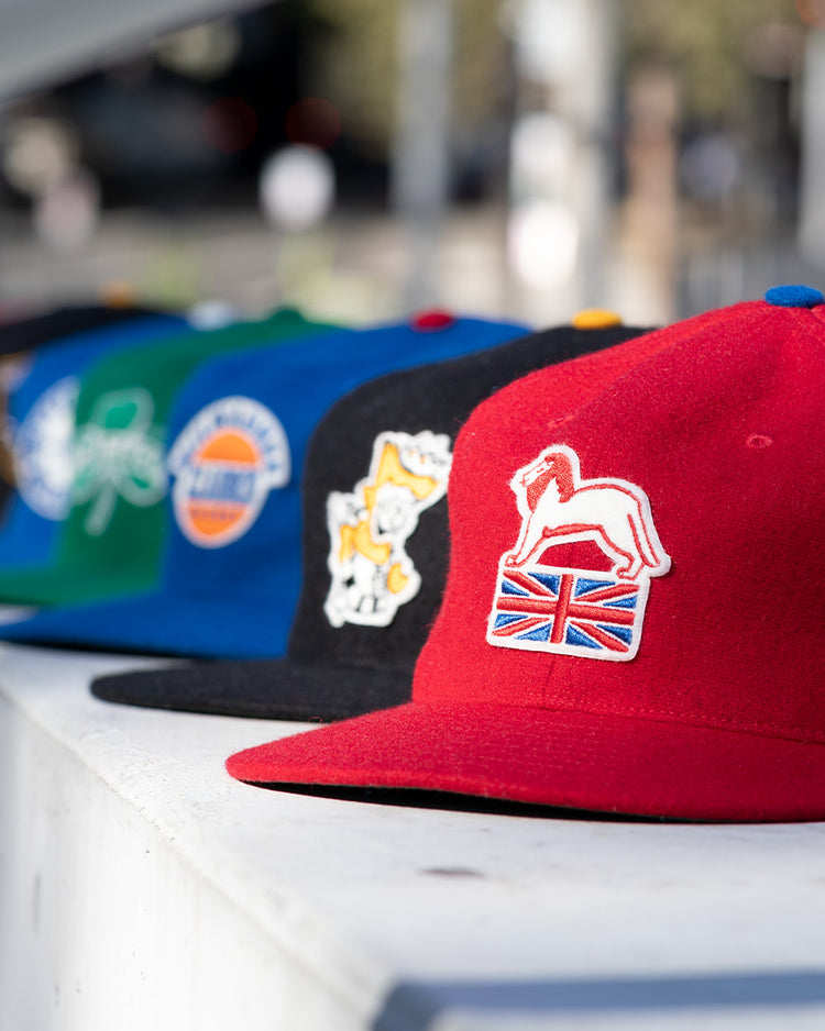 A row of colorful baseball caps is displayed outdoors, highlighting the EFF Wembley Lions 1957 Vintage Ballcap—a hand-sewn wool red cap with a white horse and British flag patch—reminiscent of classic vintage styles.