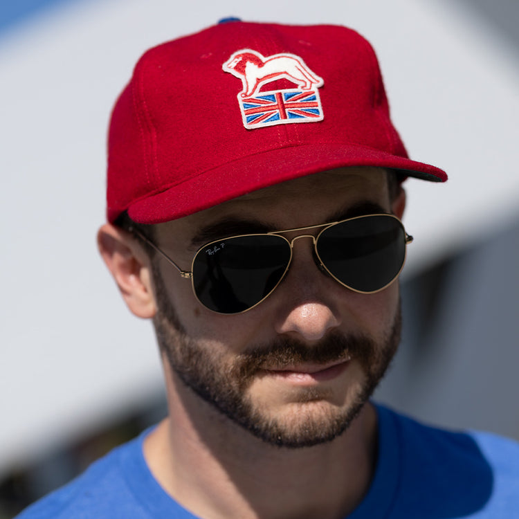 A bearded man in aviator sunglasses and an EFF Wembley Lions 1957 Vintage Ballcap with a hand-sewn white lion and Union Jack emblem, wearing a blue shirt, stands outdoors—showcasing classic British hockey pride.