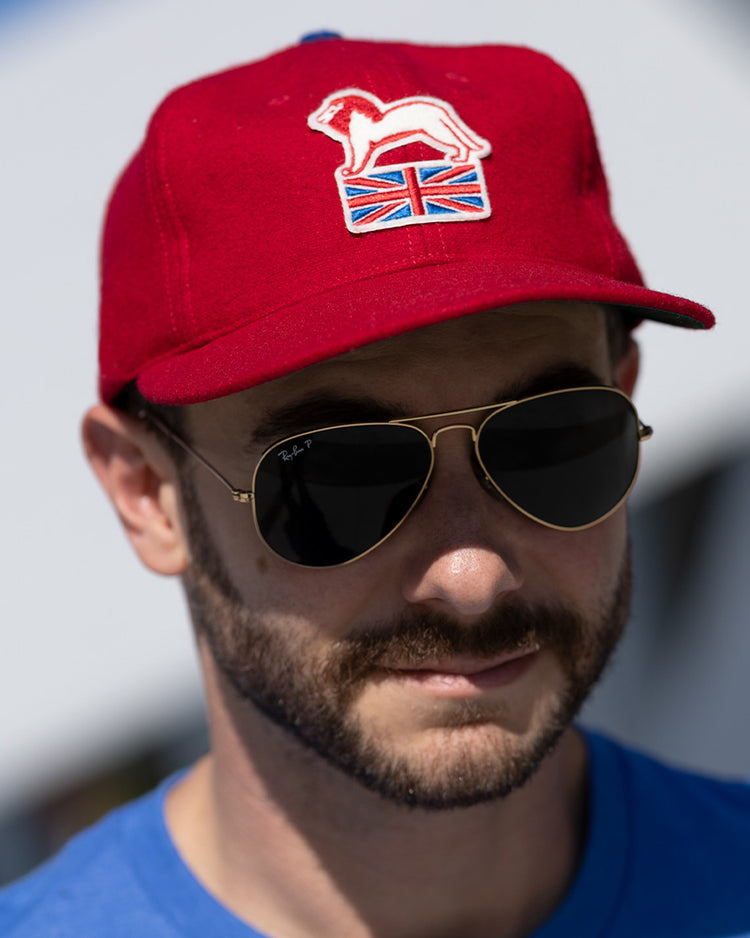 A bearded man in aviator sunglasses and an EFF Wembley Lions 1957 Vintage Ballcap with a hand-sewn white lion and Union Jack emblem, wearing a blue shirt, stands outdoors—showcasing classic British hockey pride.